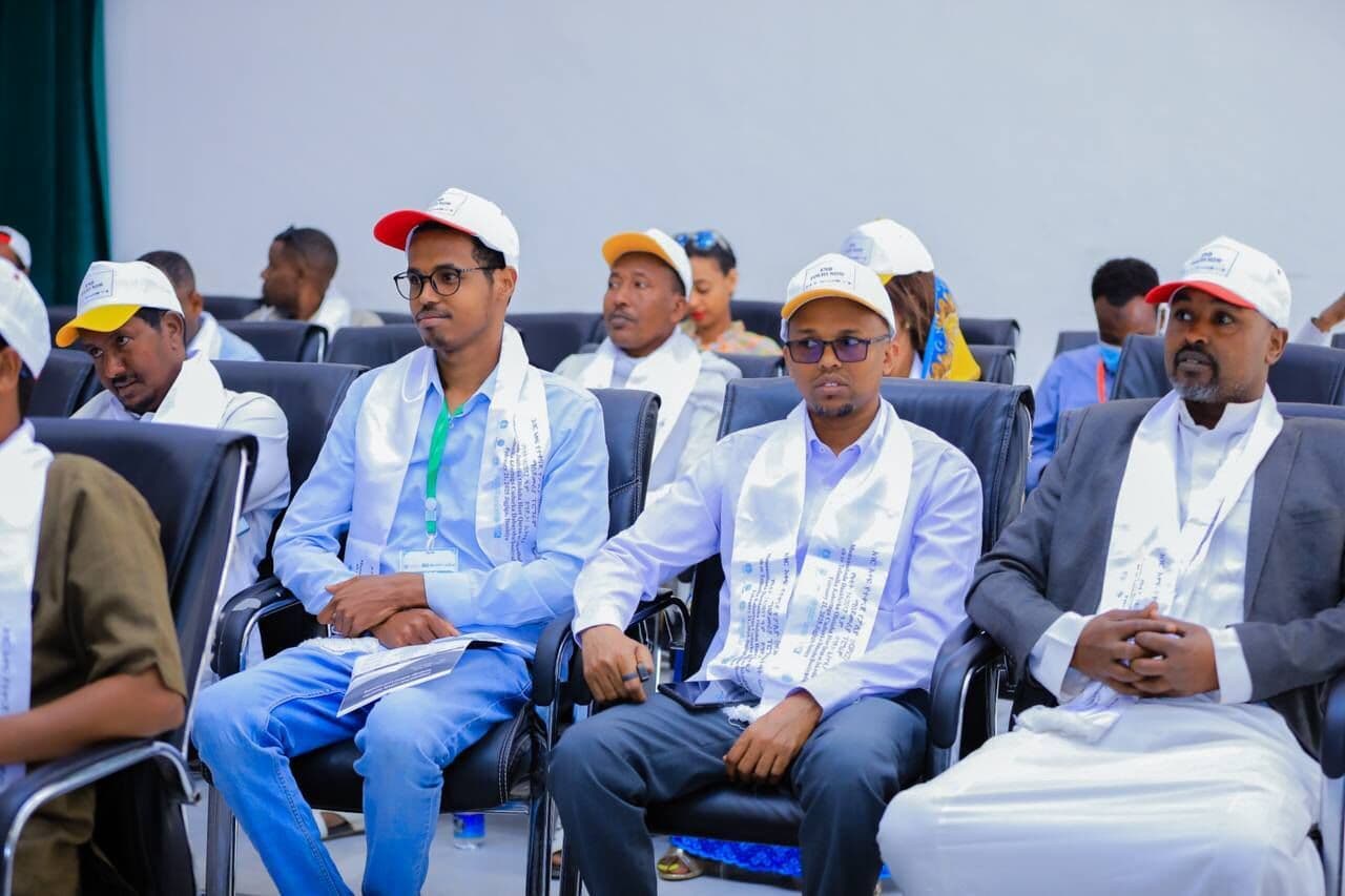 SHIFA representatives seated at a formal community health ceremony, wearing event sashes and caps